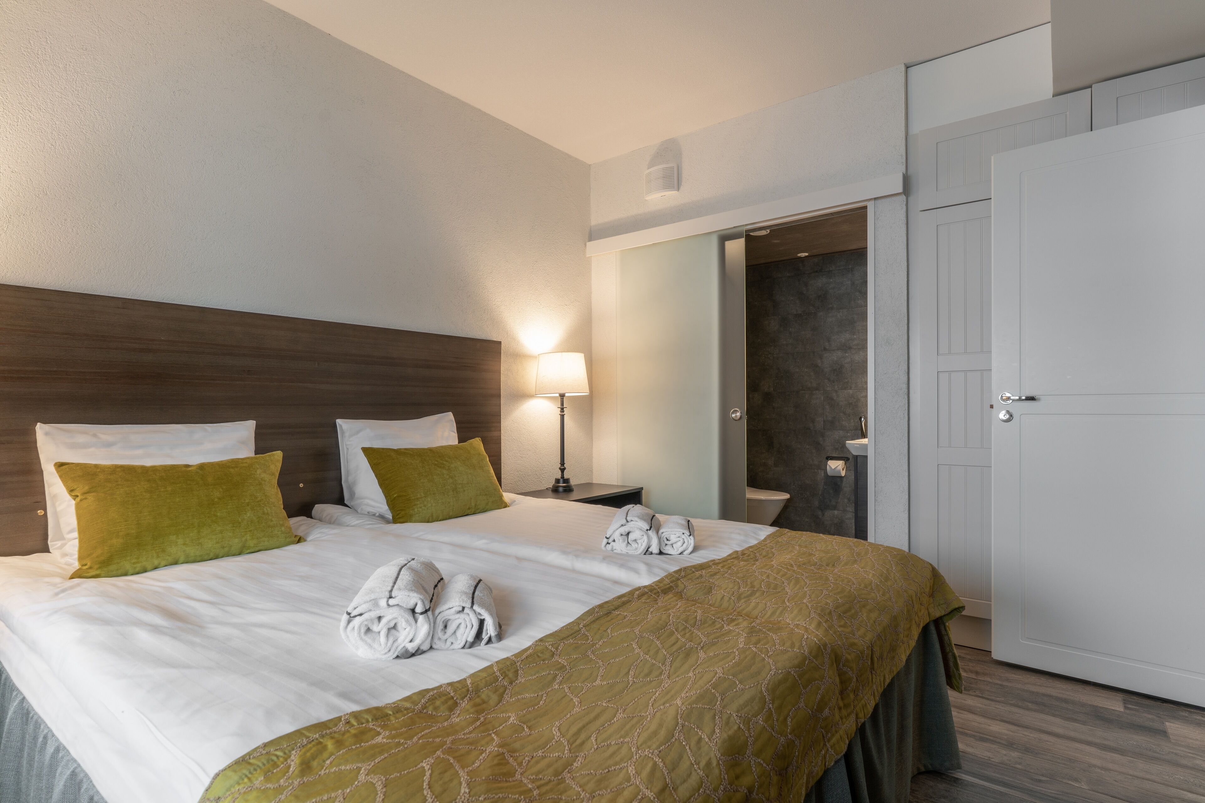 A hotel room with a large bed, two pillows, a bedside table with a lamp, a bathroom visible through an open door, and a wardrobe.