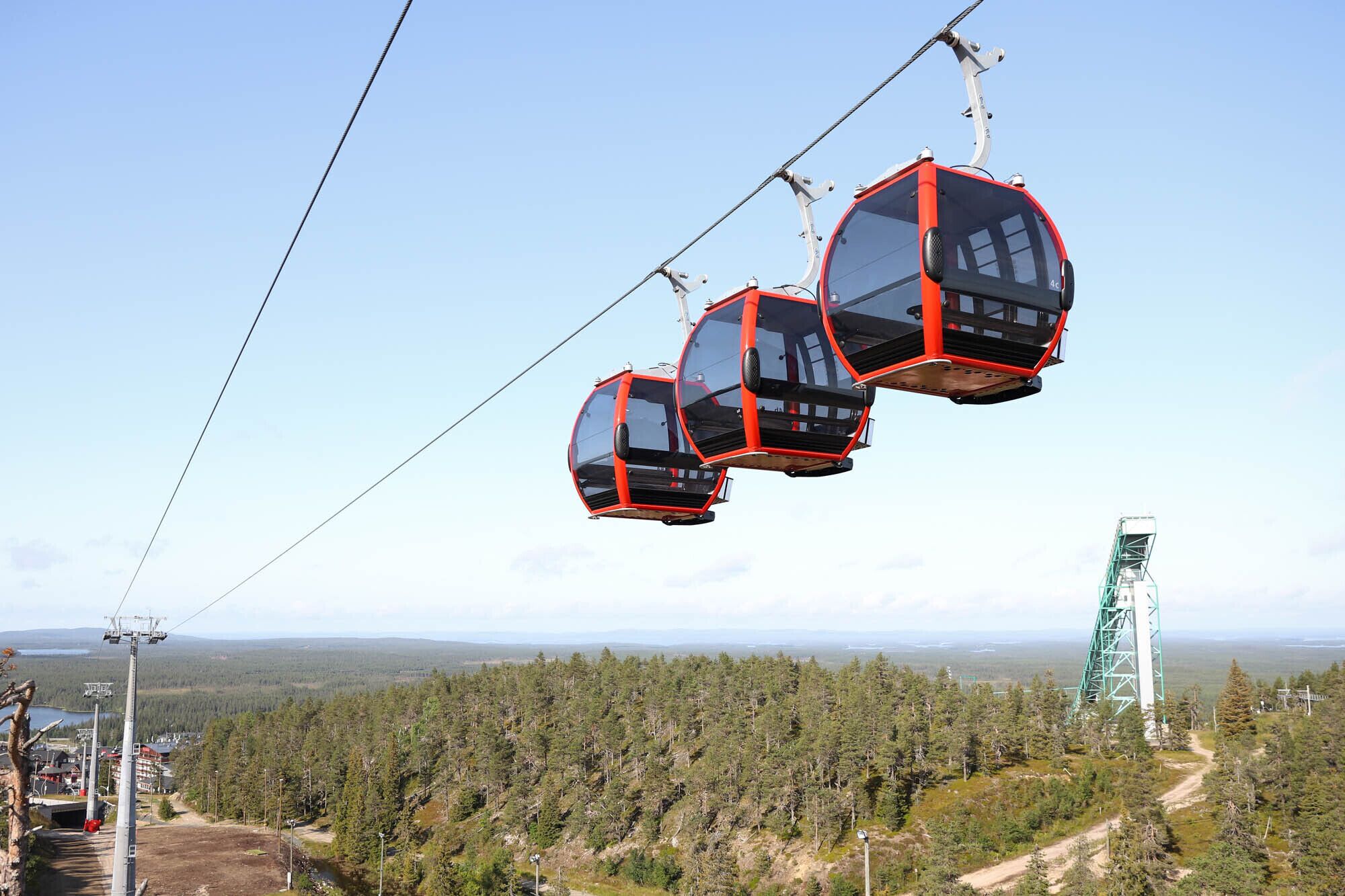 A cable car system with three cabins suspended on cables, a forested landscape below, and a distant view of a body of water and mountains.
