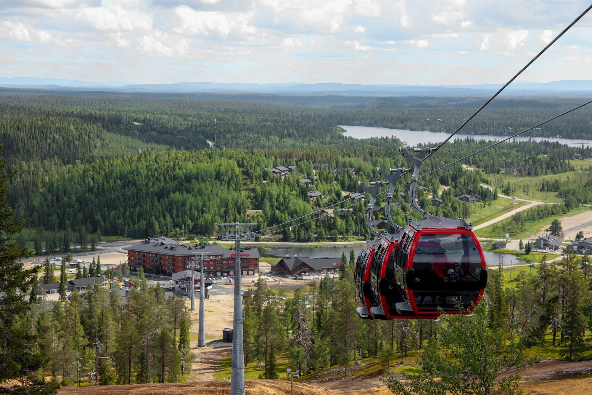 Un teleférico avanza sobre un área boscosa con un lago y edificios al fondo.
