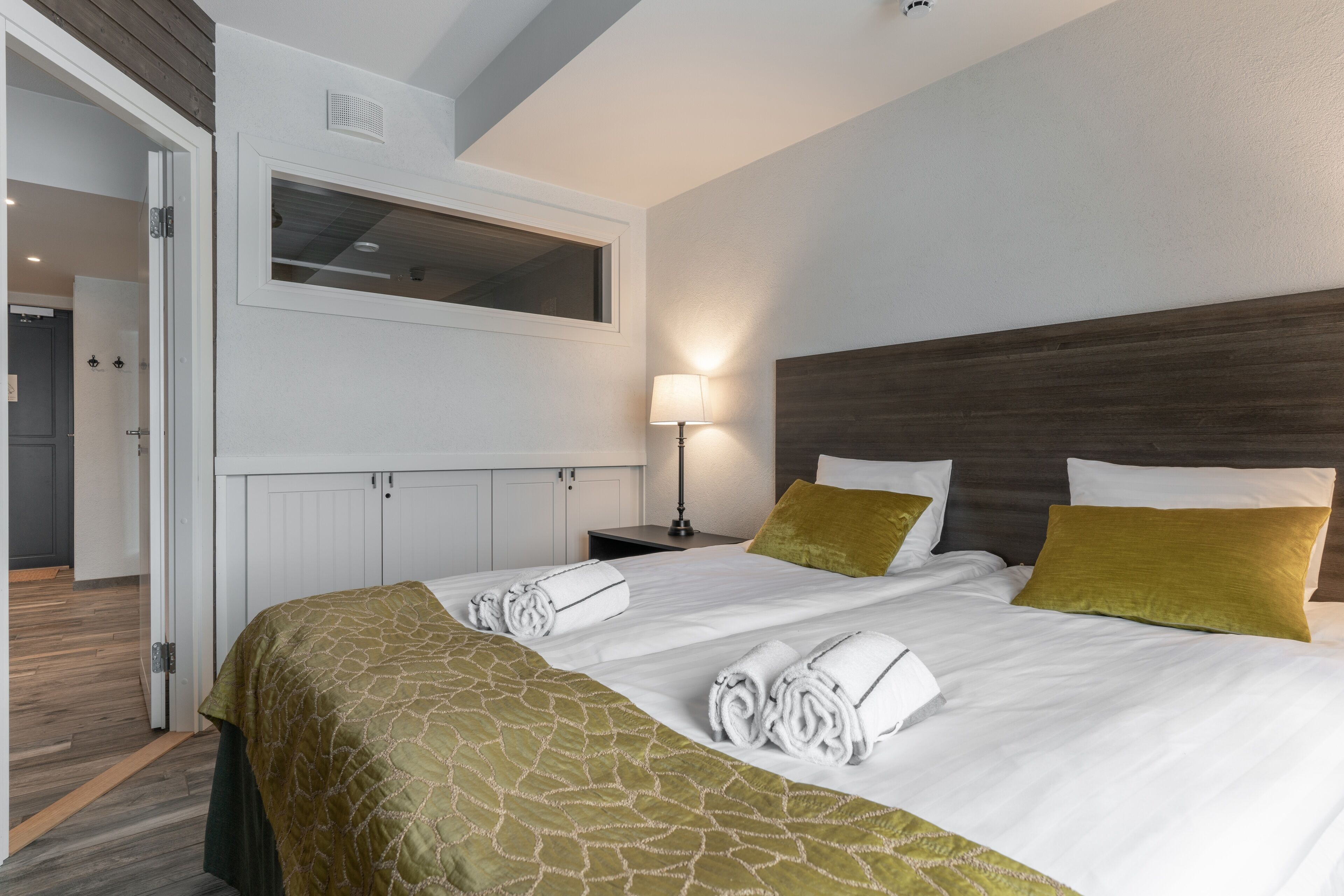 A neatly made bed with a green patterned bedspread, two pillows, and rolled towels on a white headboard.