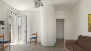 A modern living room with a beige sofa, a wooden chair, and a blue cushioned chair. A large white pillar is in the center, and a door leads to another room.