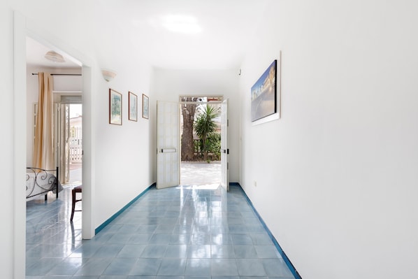 Un pasillo con suelo de azulejos azules, paredes blancas y una puerta que da a un área exterior con un árbol y vistas de un edificio.