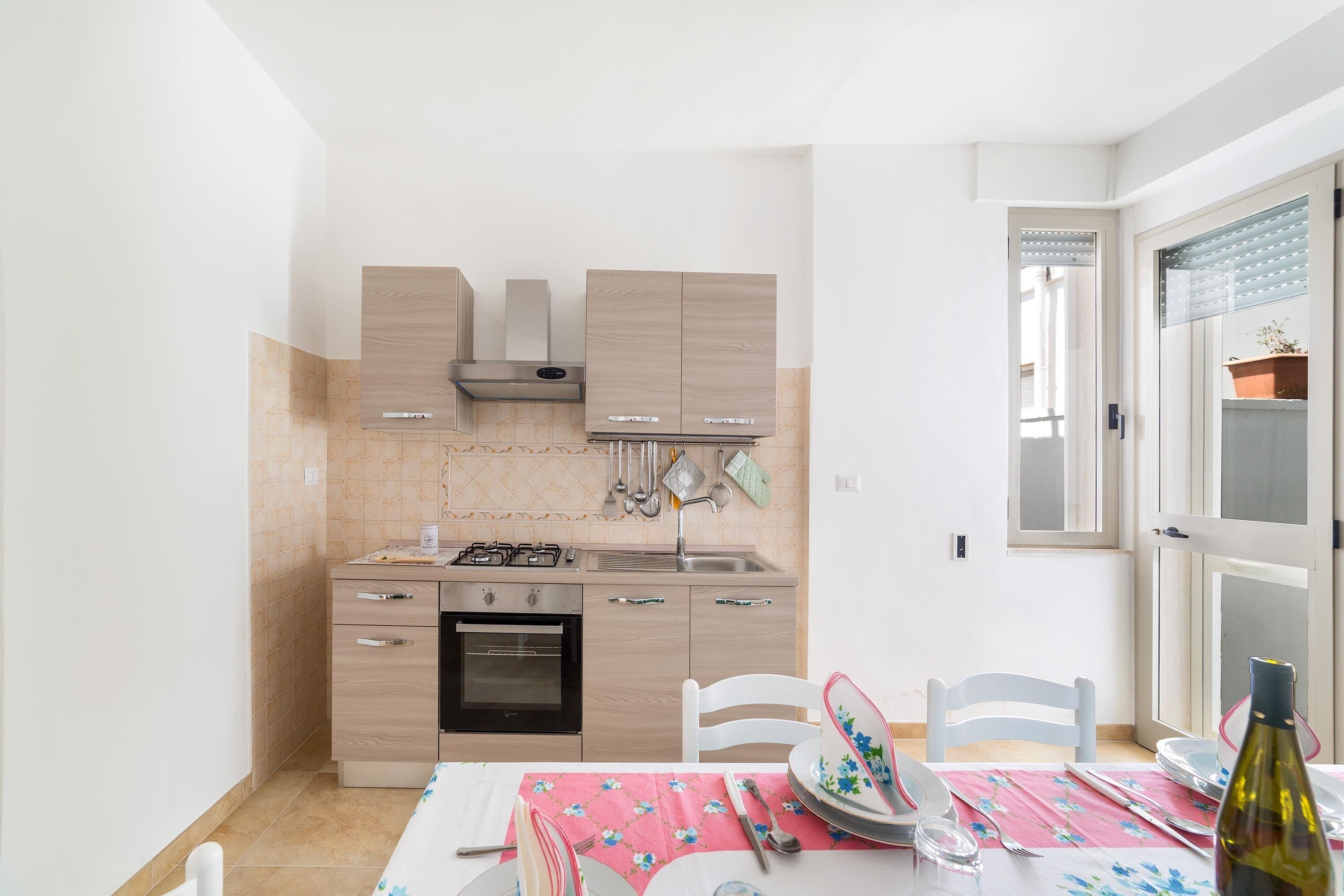 A compact kitchen with a gas stove, oven, and wooden cabinets. There is a dining table with chairs and a pink tablecloth.