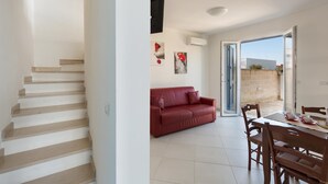 A modern living room with a staircase, a red leather sofa, a dining table with chairs, and a view through an open door. - Villetta Raffaele (Nardò)