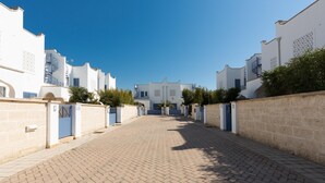 A paved street leading to a row of white buildings with blue doors and windows, surrounded by greenery. - Villetta Raffaele (Nardò)