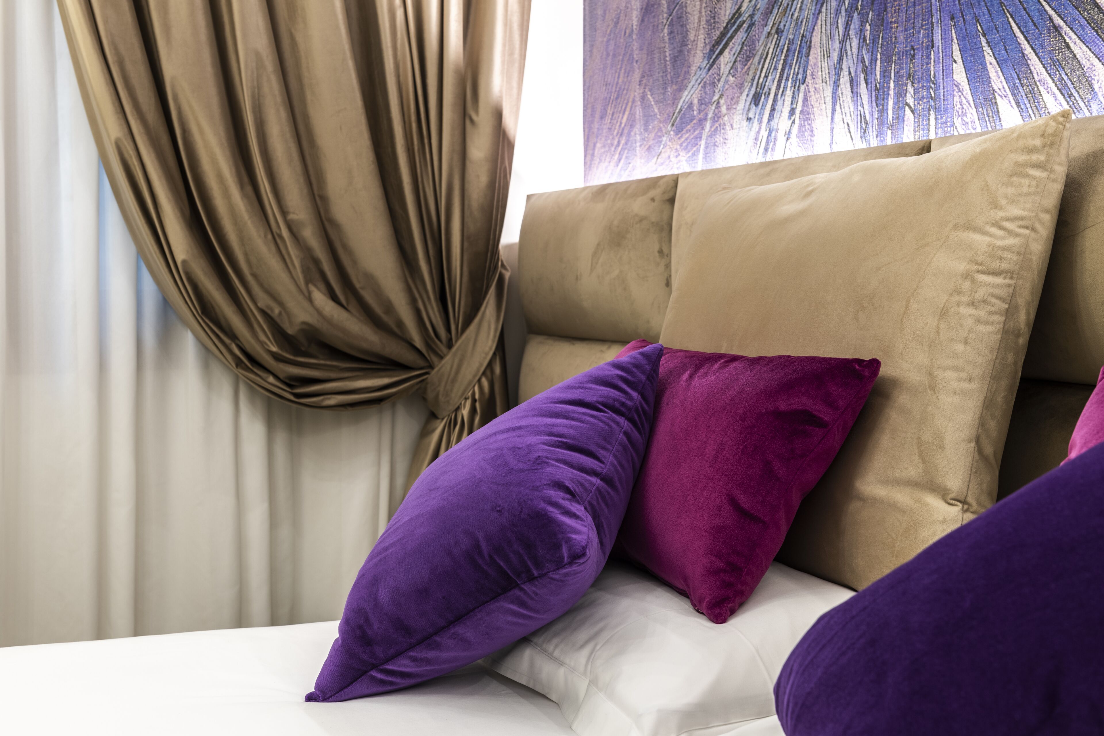 A bed with a beige headboard and multiple pillows in shades of purple and beige.