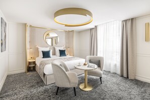 A modern hotel room with a bed, two armchairs, a round table, a mirror, and a circular light fixture.