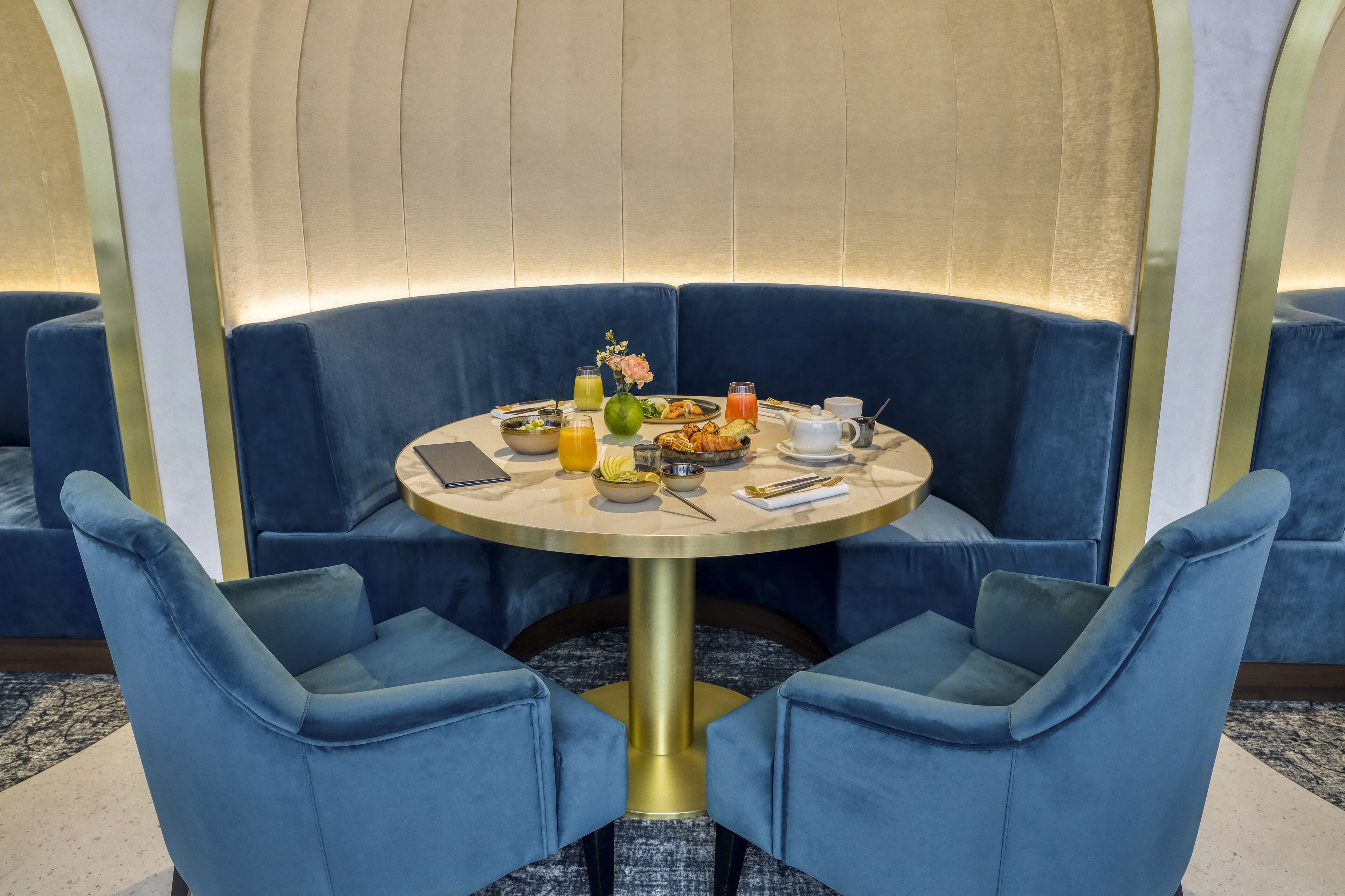 Una mesa redonda con base dorada rodeada de sillas tapizadas en azul, ubicada en una habitación con una pared curva y una alfombra estampada.