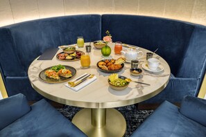 A round dining table with a marble top surrounded by blue upholstered chairs. The table is set with various breakfast items, including fruits, pastries, and beverages.