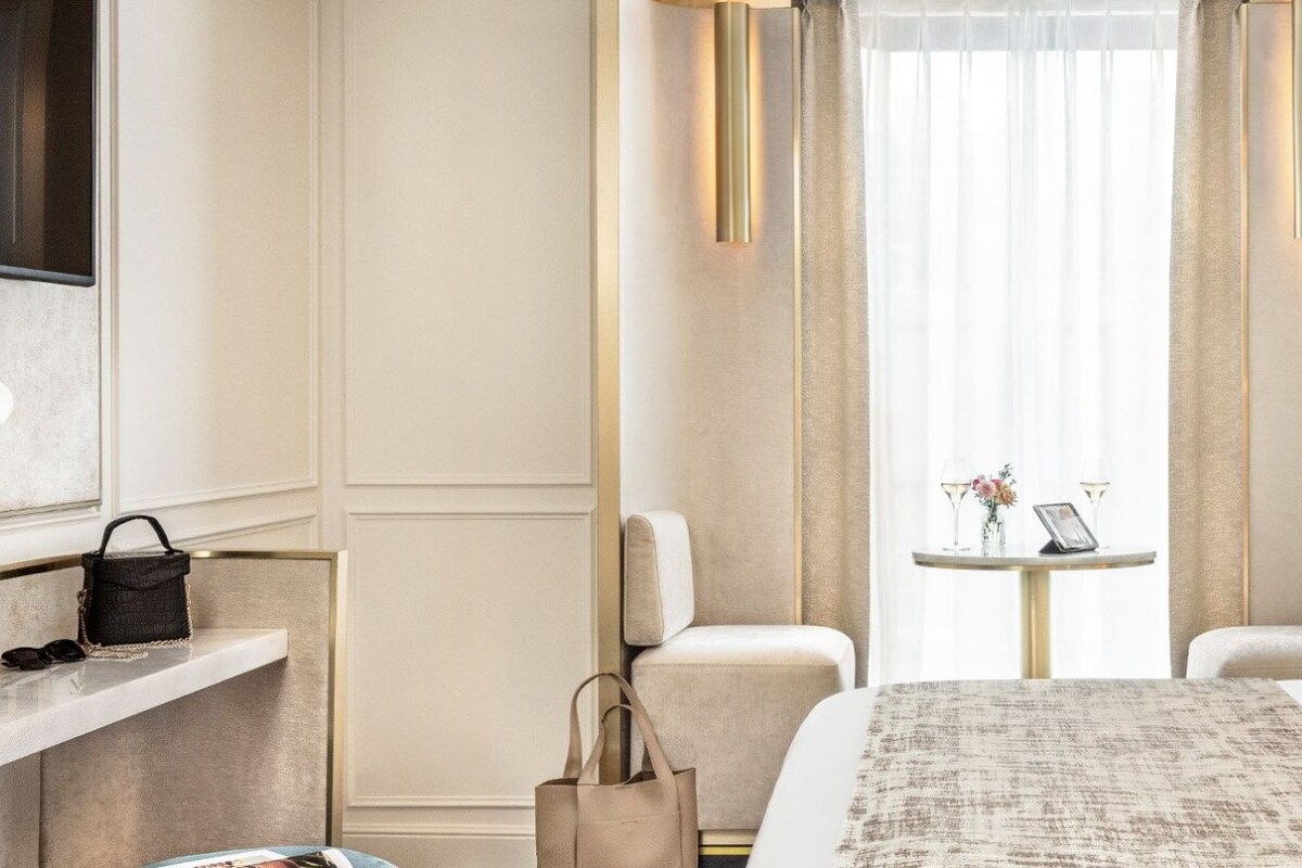 A hotel room with a bed, bedside table, chairs, a small table with a phone and glasses, and a window with curtains.