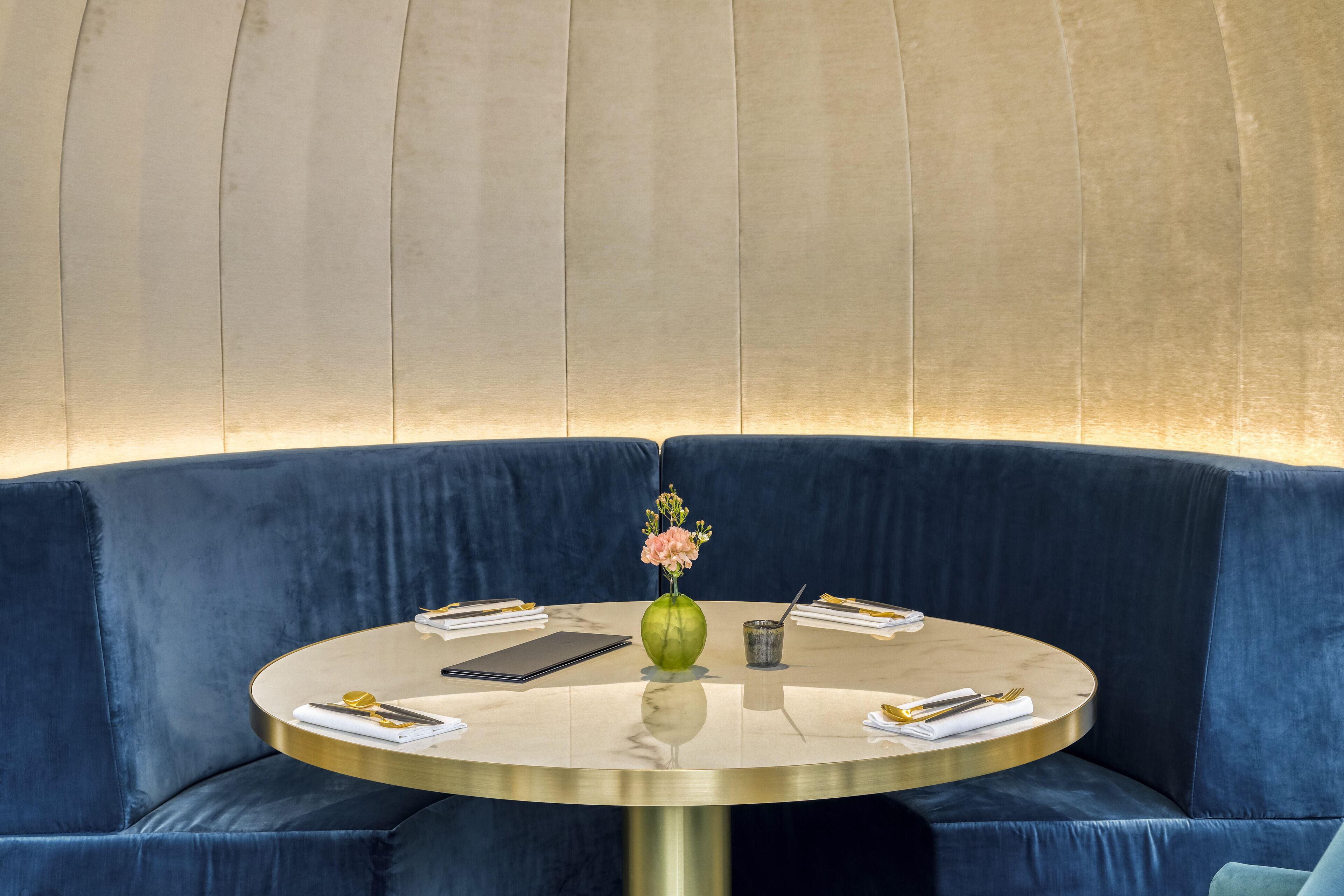 A round table with a vase of flowers, a book, and a cup, set on a blue velvet sofa against a textured wall.