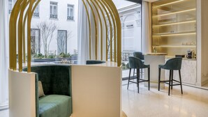 A modern interior with a green sofa, a bar area with stools, and a view of a building outside. - Maison Albar Hotels Le Vendome (Paris)
