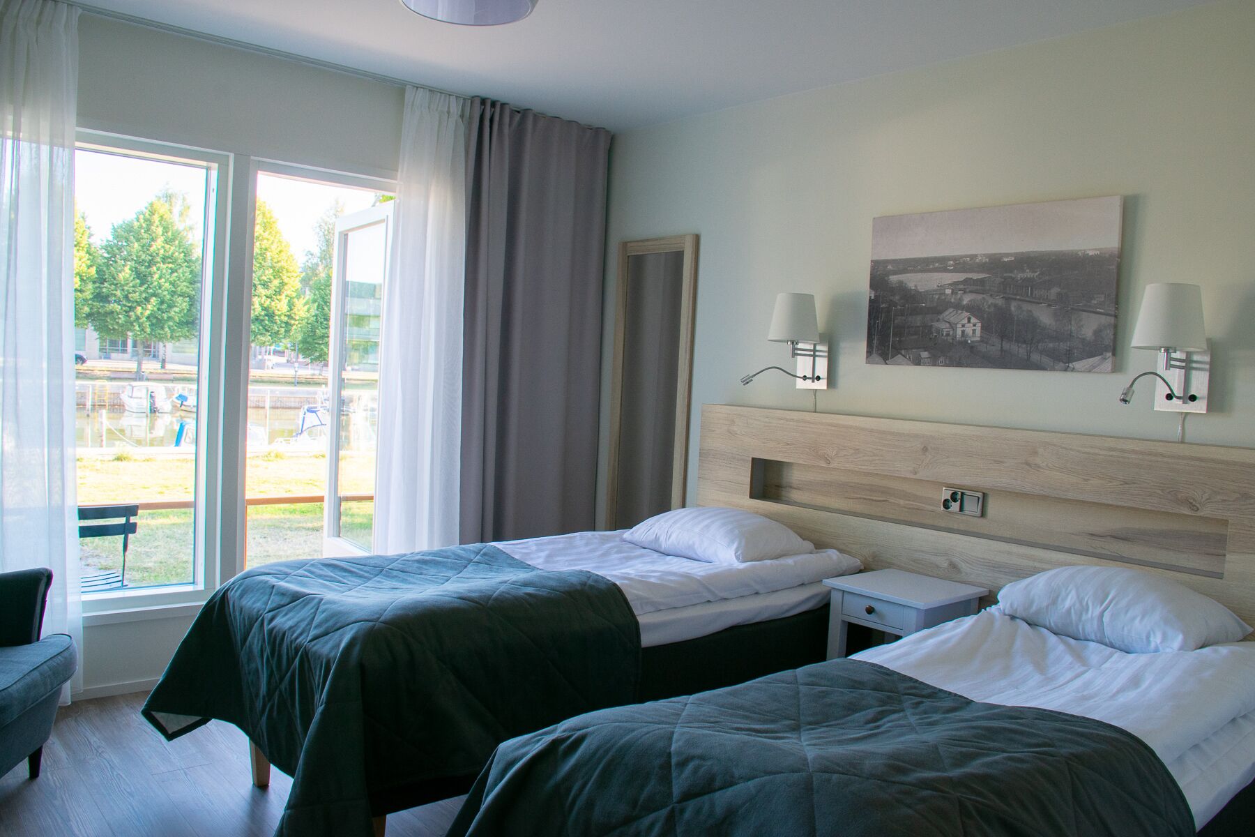 Habitación de hotel con dos camas, una mesita de noche pequeña, una silla y un ventanal grande con cortinas.