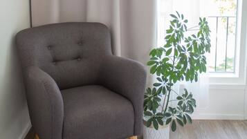 A corner room with a grey armchair, a white potted plant, and a window with sheer curtains.