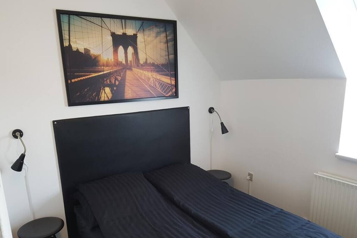 A bedroom with a bed, a wall-mounted picture, a desk with a chair, and a window.