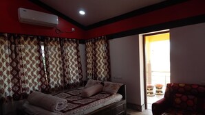 A room with a sofa bed, patterned curtains, a small table, and a view through an open door. - Asian Grand Villa (Bolpur)