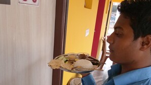 A person holding a tray with various dishes, including rice, curry, and fried items, in a room with a yellow wall and a door marked "S-3". - Asian Grand Villa (Bolpur)