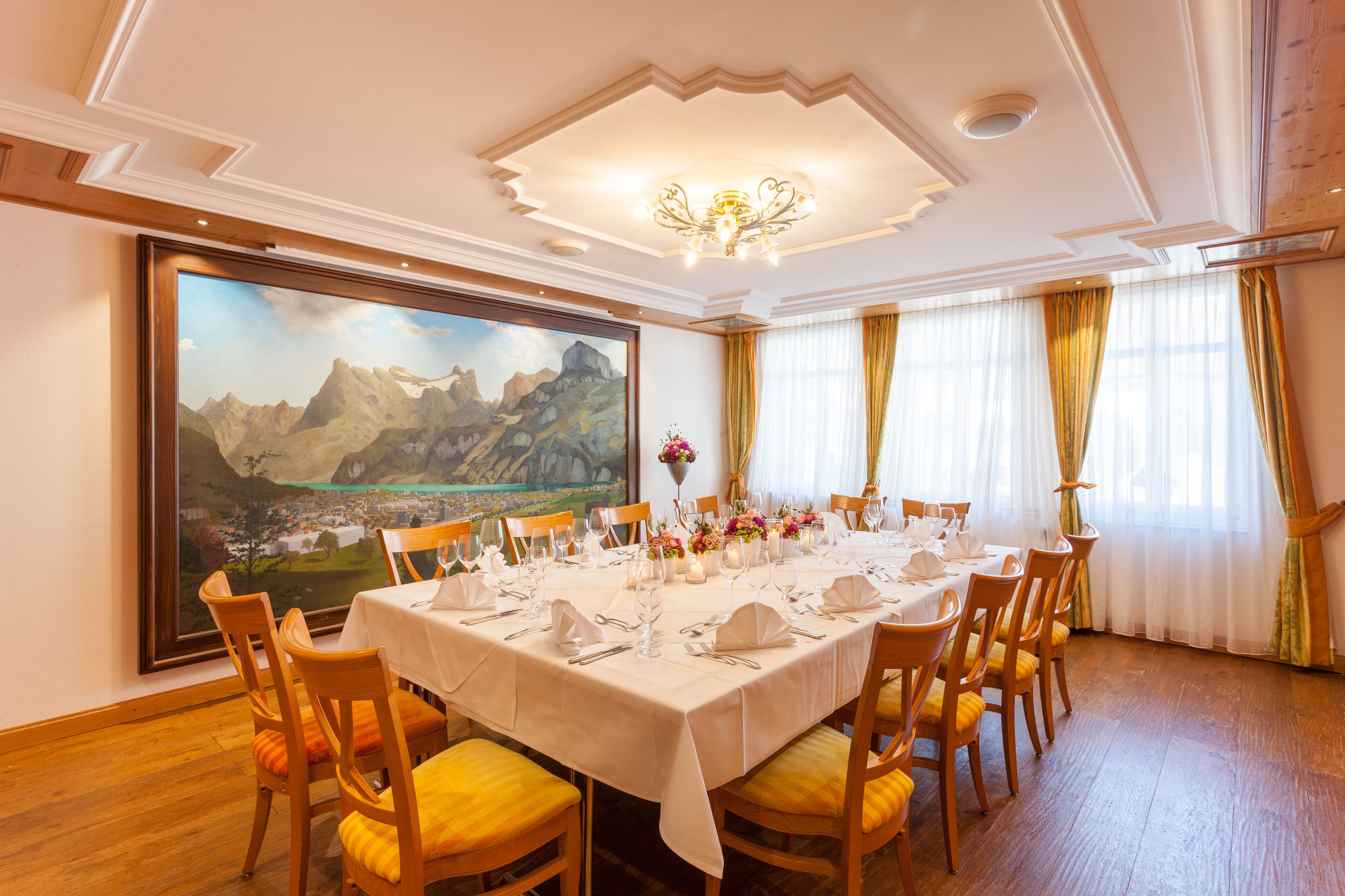 A dining room with a large table set for a meal, wooden chairs, a chandelier, and a scenic painting on the wall.