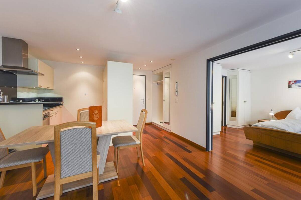 A modern, well-lit room with a wooden dining table and chairs, a kitchenette, and a bed in the adjacent room.
