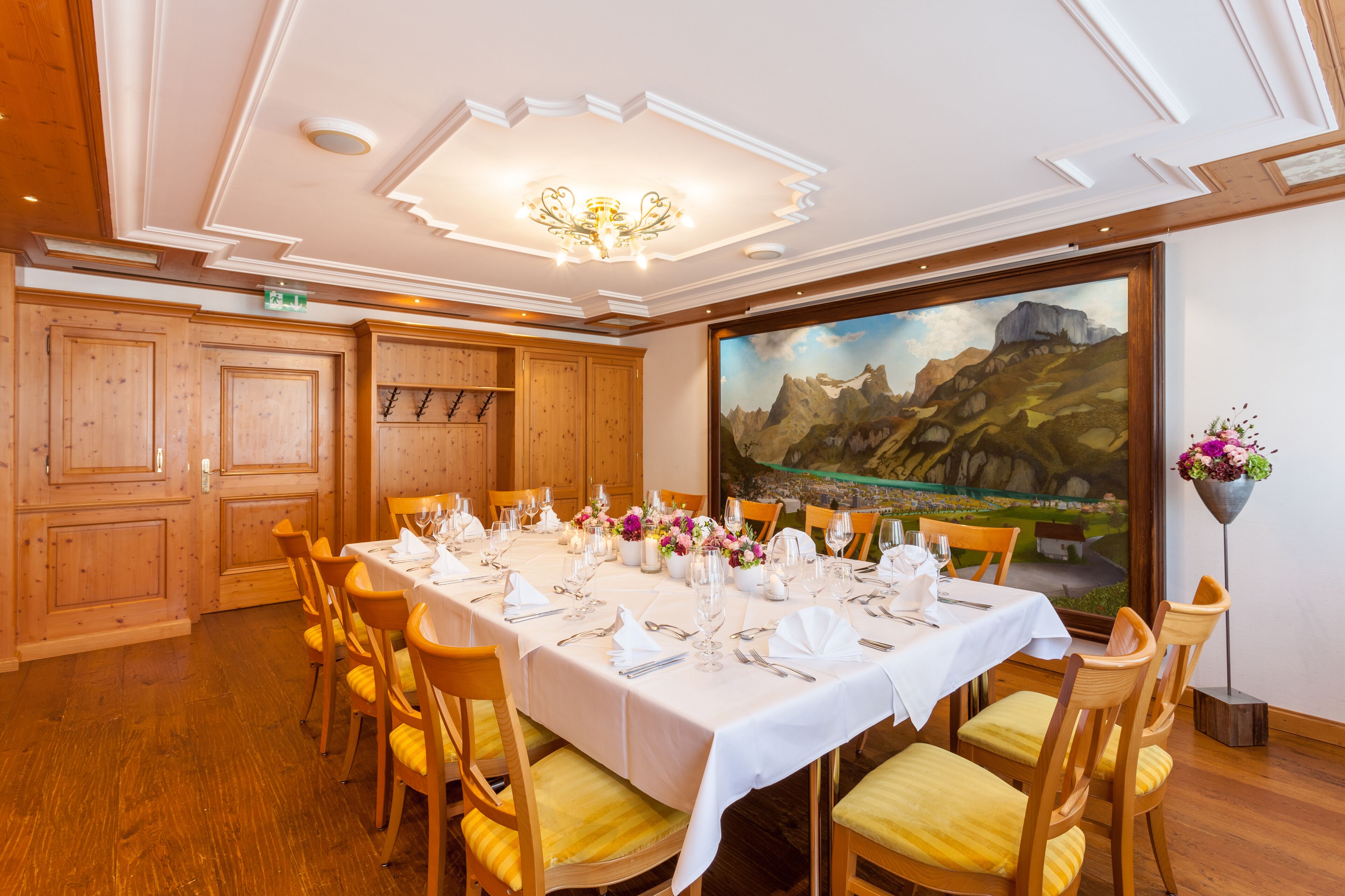 A dining room with a long table set for a meal, wooden paneling, and a large landscape painting on the wall.