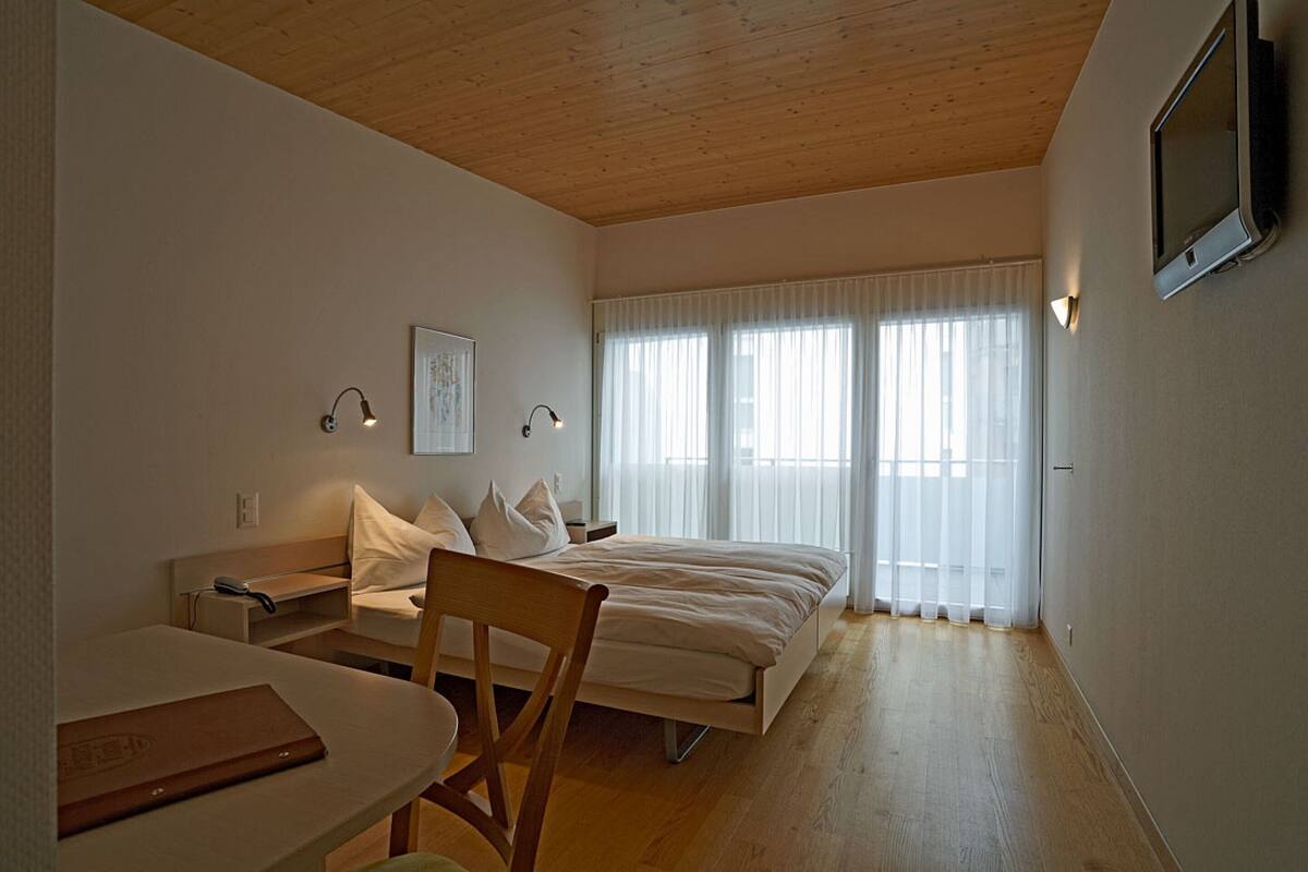 A bedroom with a wooden ceiling, a bed with white bedding, a bedside table, a chair, and a window with sheer curtains.