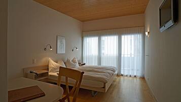 Une chambre avec un plafond en bois, un lit avec une literie blanche, une table de chevet, une chaise et une fenêtre ornée de rideaux légers.