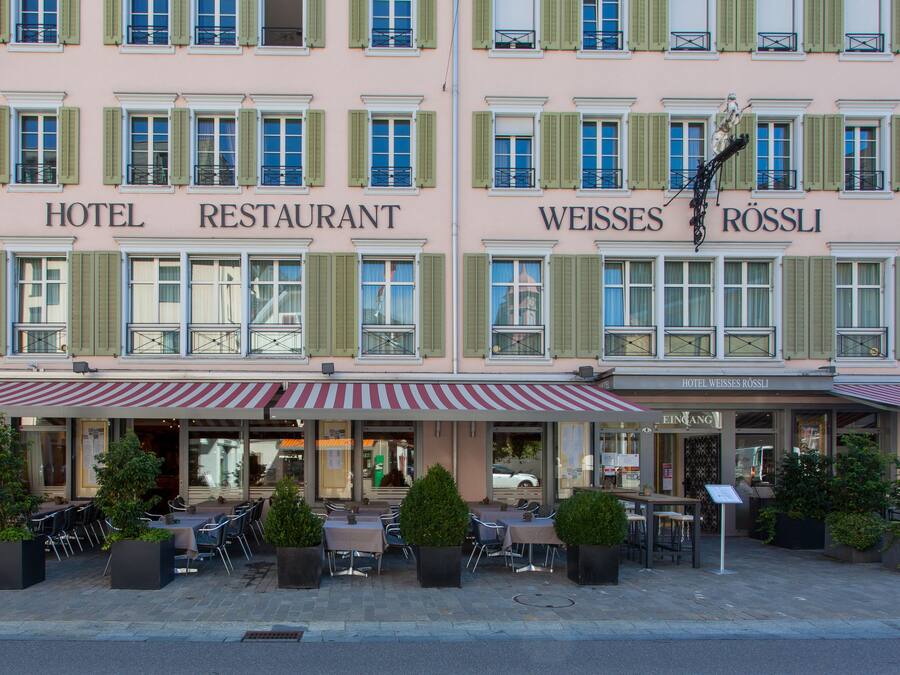 Ein Hotel mit Restaurant im Erdgeschoss, mit Außenbereich, Topfpflanzen und einem Streifenmarkisenüberdachung.