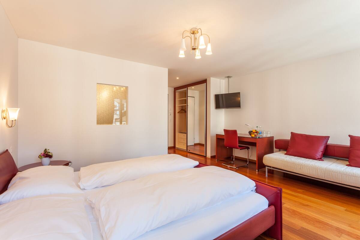 A hotel room with a large bed, a sofa, a desk with a chair, a TV, and a mirror.