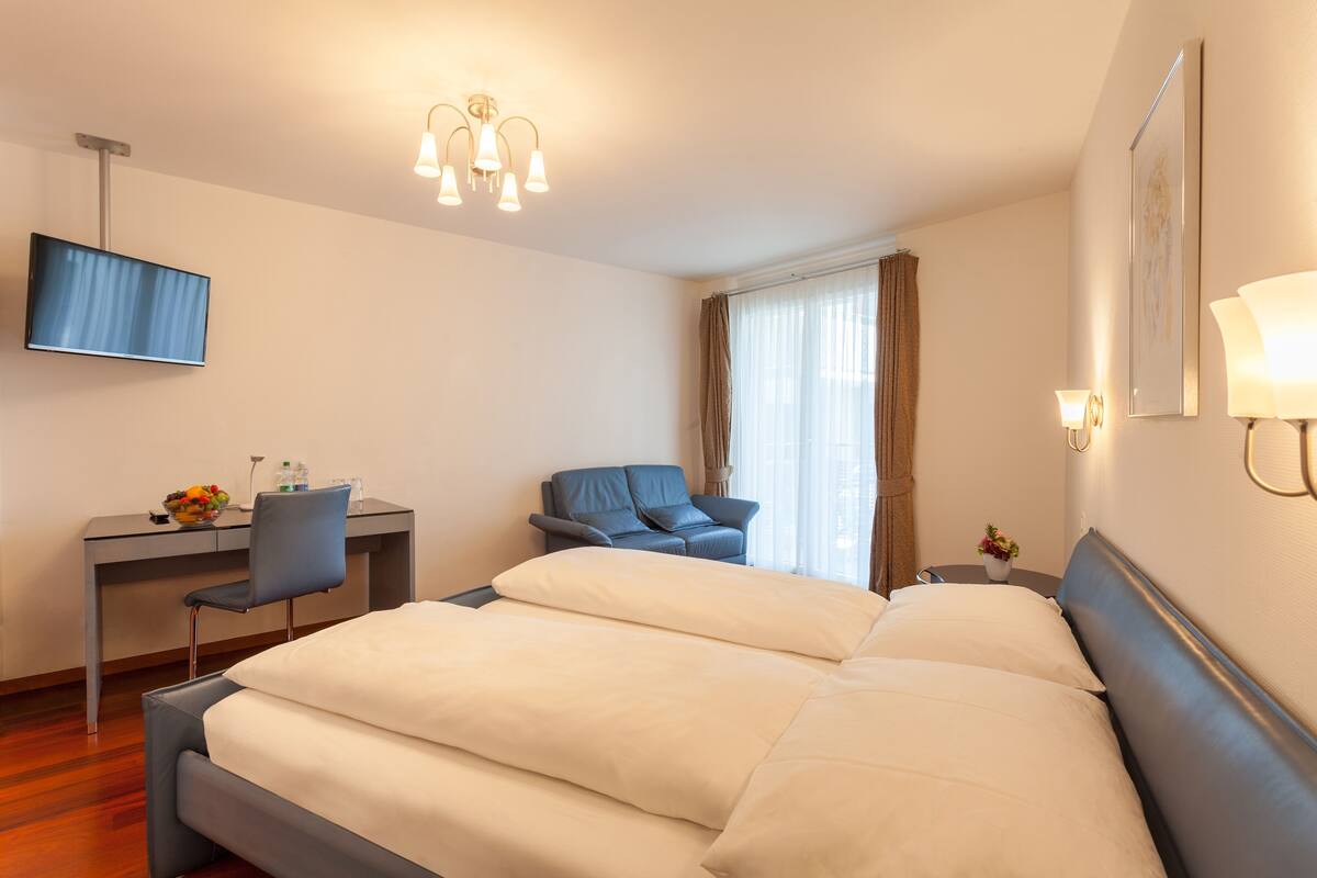 A hotel room with a large bed, a desk with a chair, a flat-screen TV, and a small table with a lamp.