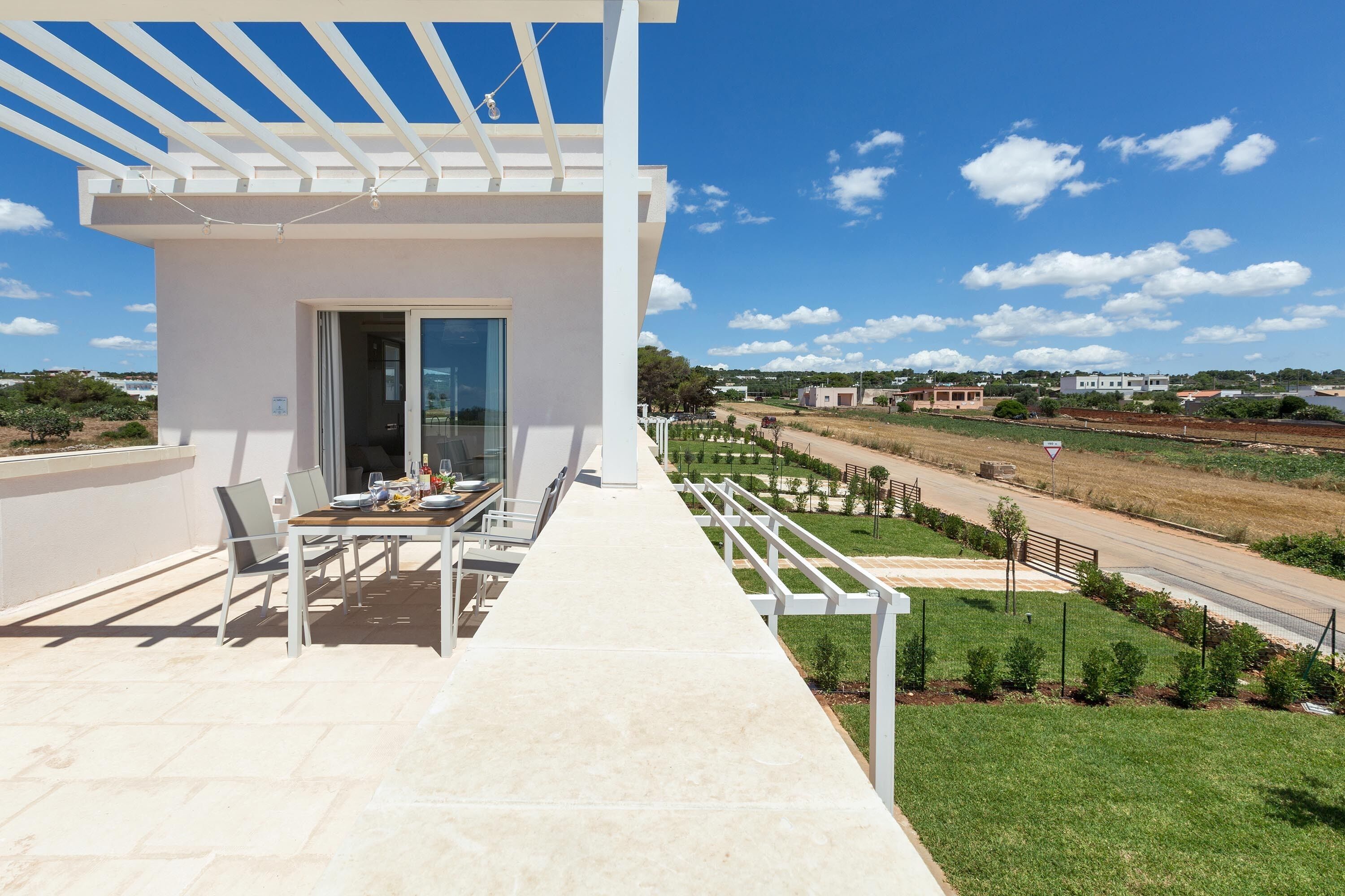 A modern outdoor patio with a table and chairs, a pergola, and a view of a green landscape and distant buildings.