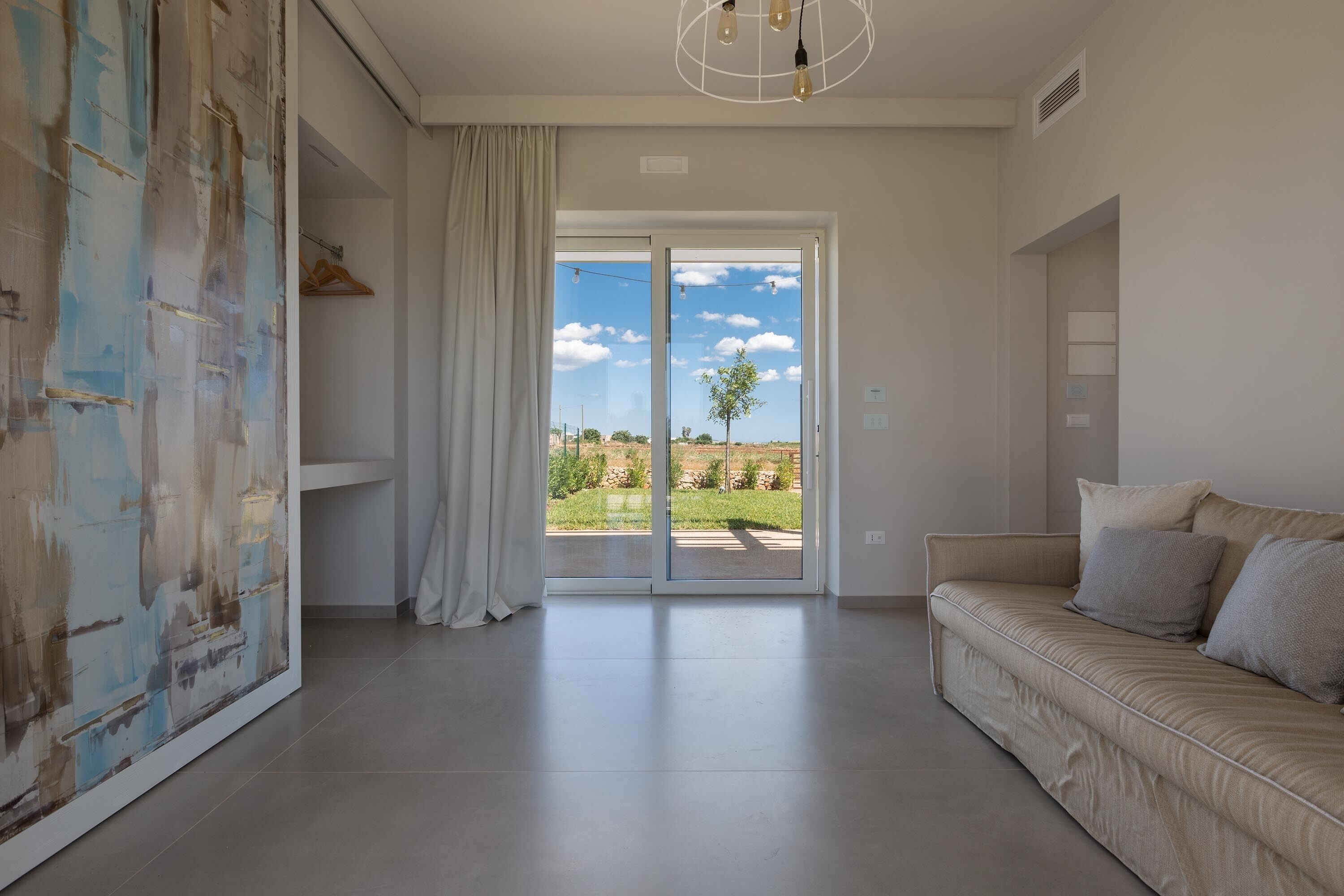 A modern living room with a beige sofa, a large abstract painting, and a sliding glass door leading to a garden.