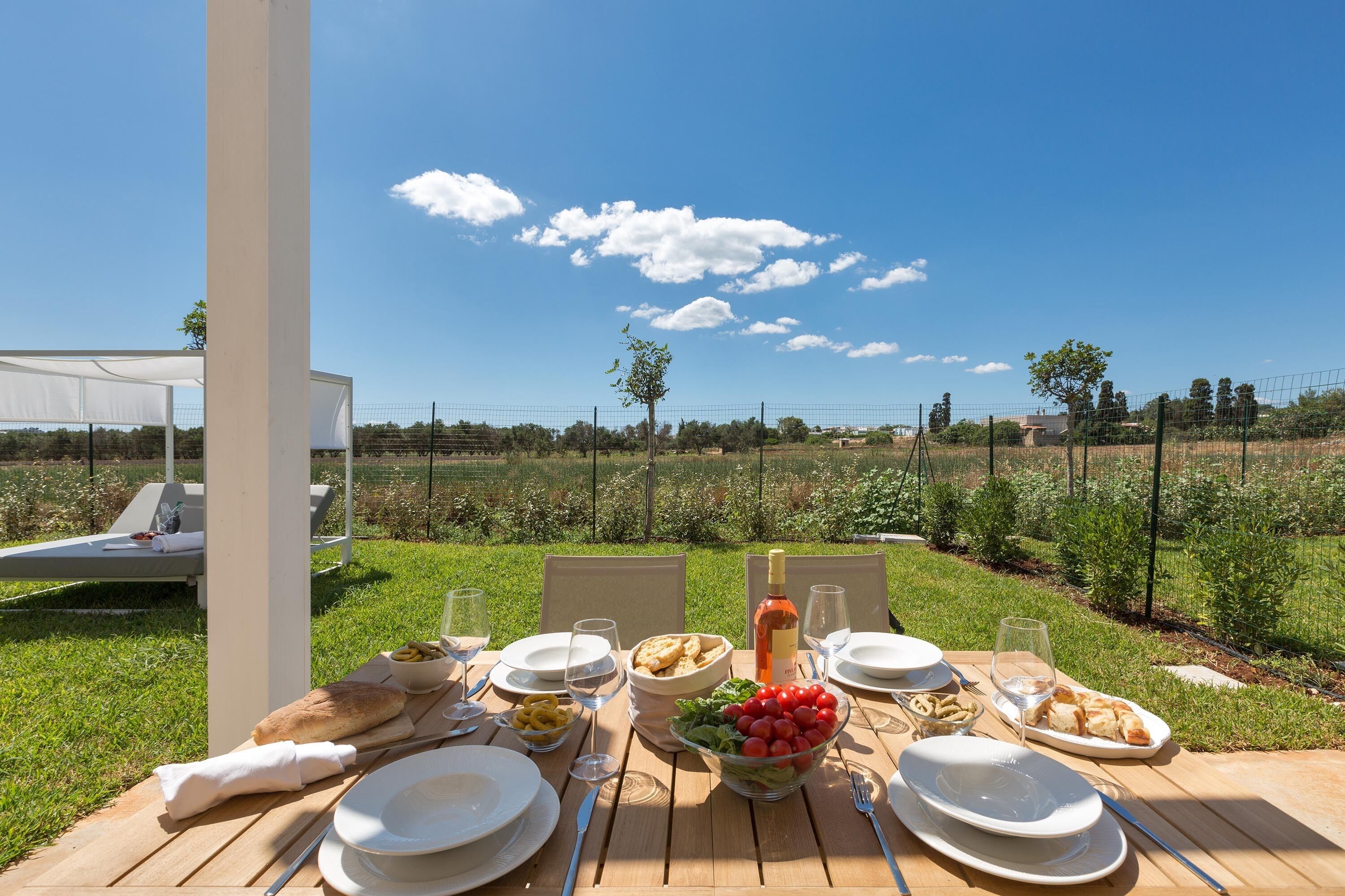 An outdoor dining table set with plates, glasses, and food, overlooking a garden and a clear sky.