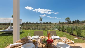 An outdoor dining table set with plates, glasses, and food, overlooking a garden and a clear sky.