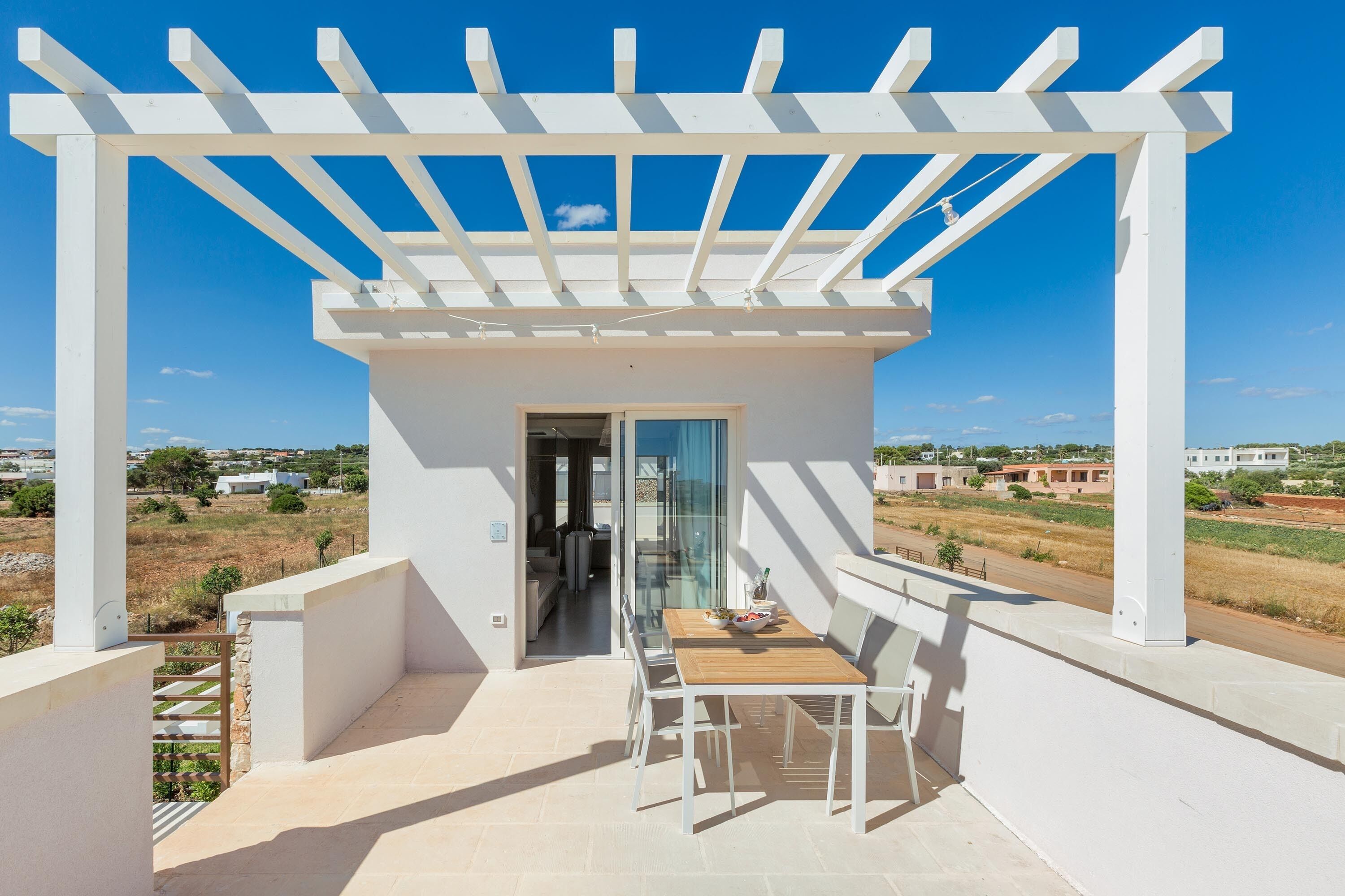 A balcony with a pergola, a table with chairs, and a view of the surrounding landscape.