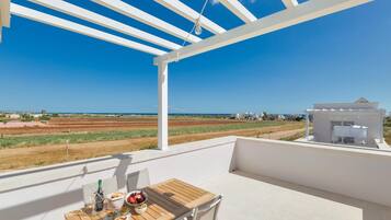Un balcon avec une table en bois, des chaises blanches et une vue sur la mer et les champs.
