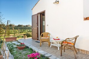 A patio with two wicker chairs and a small table, a door, and a view of a green landscape.
