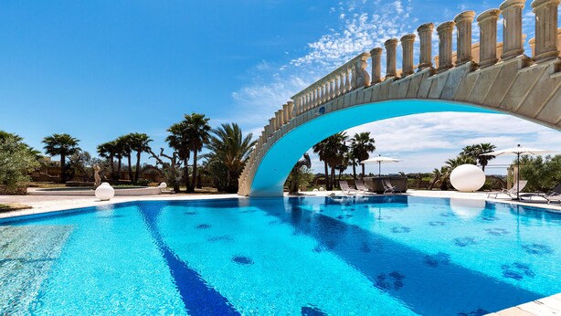 A swimming pool with a large arch bridge, palm trees, and lounge chairs. - Villa Flo (Sannicola)