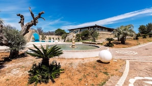 A landscaped garden with a fountain, palm trees, and a building with a blue archway. - Villa Flo (Sannicola)