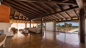 A spacious living area with wooden flooring, a high ceiling, and large windows offering a view of palm trees and a bridge. - Villa Flo (Sannicola)