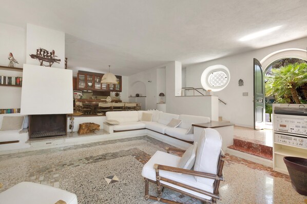 A modern living room with a white sectional sofa, a fireplace, a dining area, and a circular window overlooking the outdoors. - Villa Denise (Castellaneta)