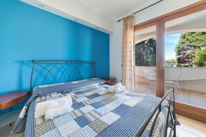 Un dormitorio con pared azul, estructura de cama metálica, colcha a cuadros y vistas a un balcón con mobiliario exterior.