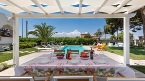 Un área de comedor al aire libre con vista a la piscina, una pérgola blanca y asientos exteriores.