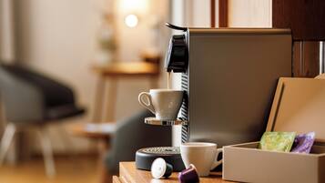 Una cafetera sirviendo café en una taza, una caja con cápsulas de café y una taza sobre una mesa de madera en una habitación con una silla y un armario.