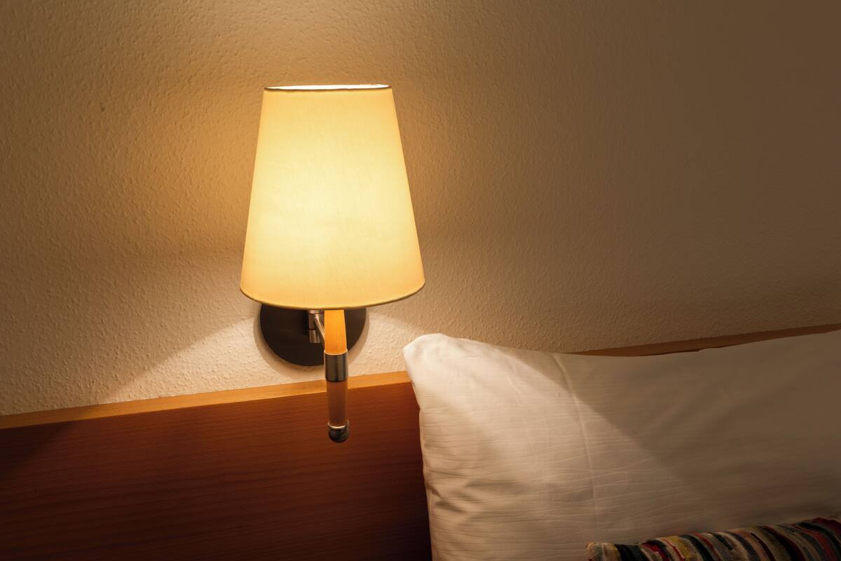 Una lámpara de pared con pantalla blanca, una mesita de noche de madera y una almohada blanca.