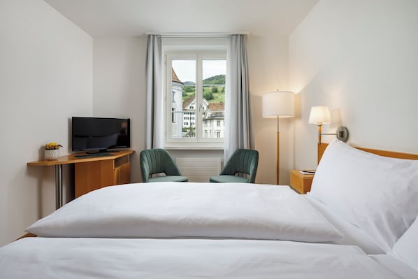 Habitación de hotel con cama, escritorio con televisor, dos sillas, ventana con vista a edificios y lámpara de noche.