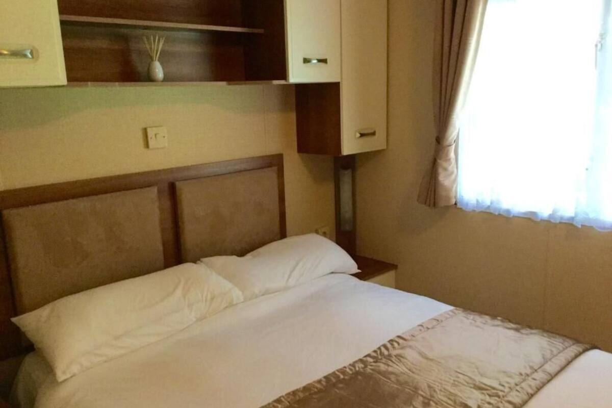 Una habitación de hotel con una cama grande, un cabecero de madera, una mesita de noche y una lámpara, junto a una ventana con cortinas.