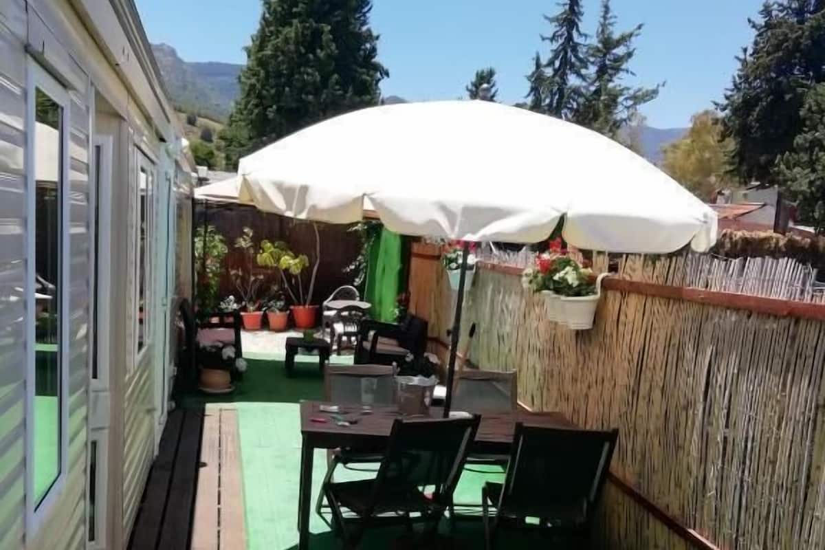 Un espacio al aire libre con terraza de madera, césped artificial verde, una mesa con sillas y un amplio paraguas blanco.