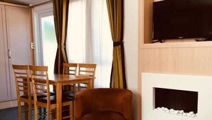 Una habitación con una mesa de comedor y sillas de madera, un sillón marrón, un televisor de pantalla plana montado en una pared de madera y una chimenea con piedras blancas.