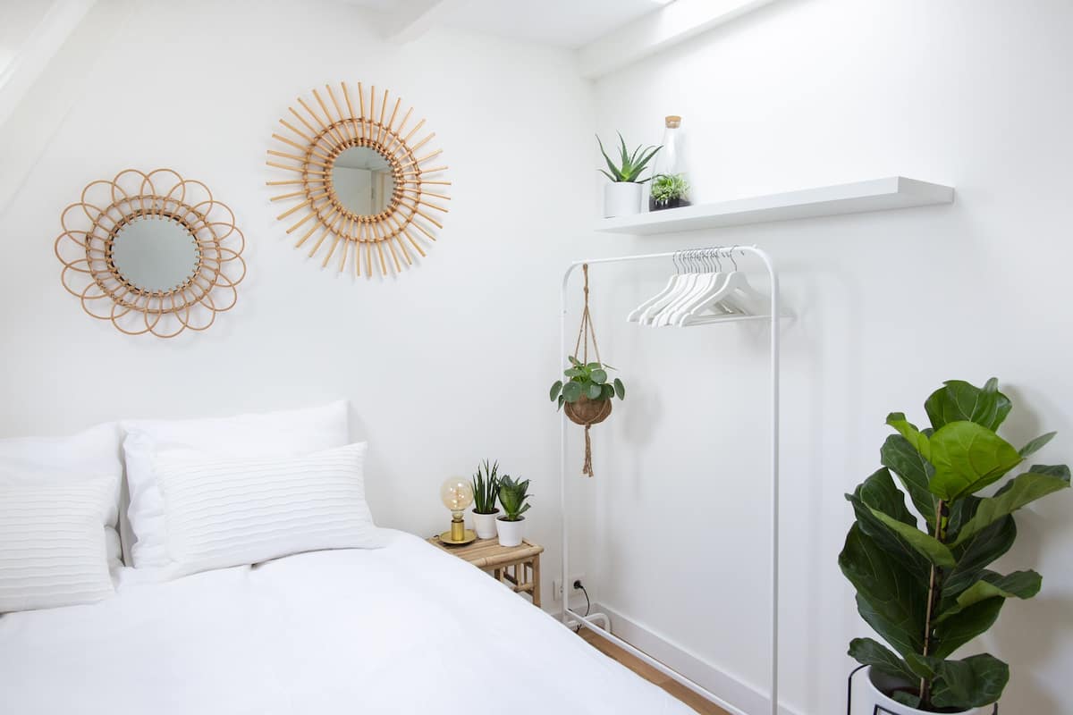 A bedroom with a bed, two wall mirrors, a hanging plant, a shelf with plants, and a wall-mounted drying rack.