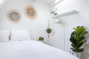 Een slaapkamer met een bed, witte beddengoed, twee spiegelwanden, een hangplant, een witte plank met een plant, en een witte kledingkast met een hangstang.